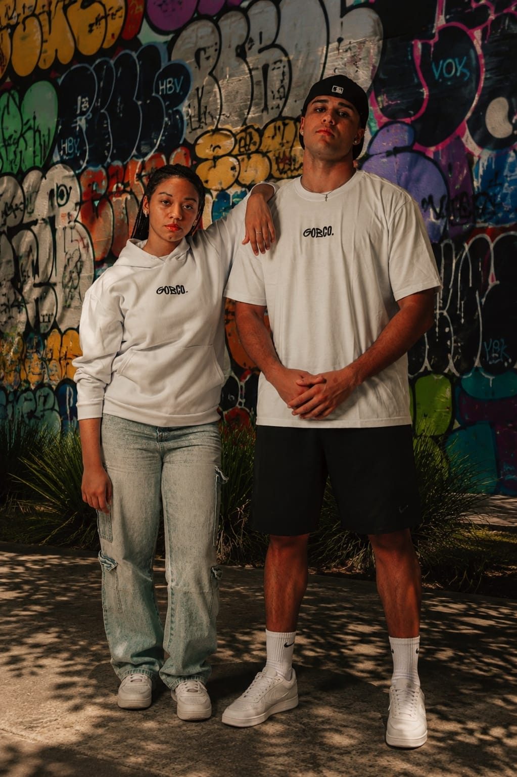 Casal vestindo camiseta e moletom GOBCO em frente a muro grafitado